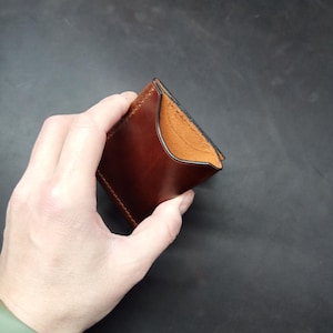 May include: A brown leather wallet with a black stitched edge. The wallet is folded in half and held in a hand.