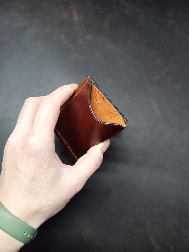 May include: A brown leather wallet with a black stitched edge. The wallet is folded in half and held in a hand.