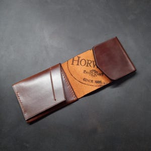 May include: A brown leather wallet with a fold-over flap. The wallet has a small pocket on the front and the Horween Leather Company logo is embossed on the inside.