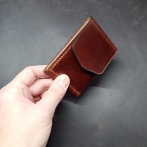 May include: A brown leather wallet with a flap closure. The wallet is held in a hand.