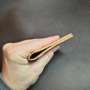 Op de afbeelding: Een lichtbruine leren portemonnee, dubbelgevouwen, in een hand gehouden. De portemonnee lijkt leeg en heeft een eenvoudig, minimalistisch ontwerp. Het leer heeft een natuurlijk, getextureerd uiterlijk.