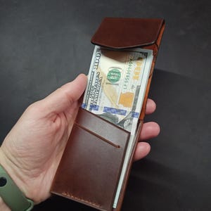 May include: A brown leather wallet with a flap closure. The wallet is open and has a billfold and a card slot. The wallet is holding a stack of US dollar bills.