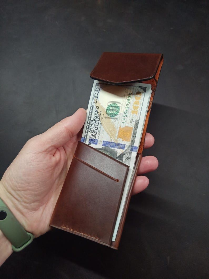May include: A brown leather wallet with a flap closure. The wallet is open and has a billfold and a card slot. The wallet is holding a stack of US dollar bills.