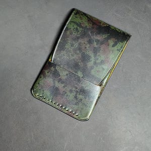 May include: A rectangular leather card holder featuring a mottled camouflage pattern in shades of dark green, black, and hints of deep red. The accessory has visible stitching along the bottom edge and a slanted top pocket opening.