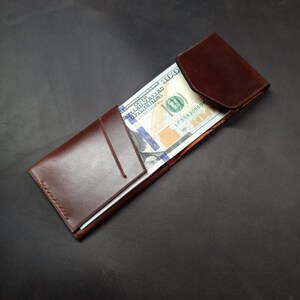 May include: A brown leather wallet with a flap closure. The wallet is open and contains a US dollar bill.