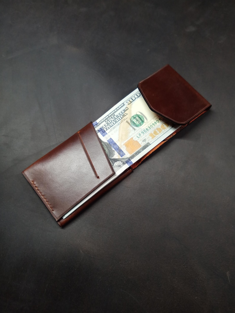 May include: A brown leather wallet with a flap closure. The wallet is open and contains a US dollar bill.