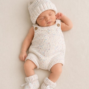 May include: A cream-colored knitted baby outfit consisting of a beanie, a romper with two small brown buttons at the shoulders, and a pair of booties. The fabric features a speckled pattern with tiny flecks of blue, pink, and yellow throughout.