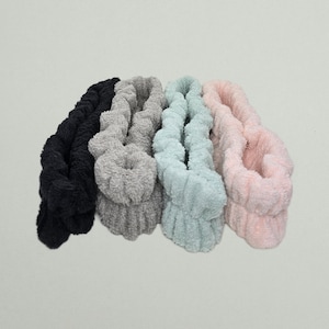 May include: Four plush headbands in a row, each a different color. The colors are black, gray, light blue, and pink. The headbands are made of a soft, textured material.