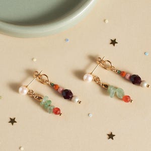 May include: A pair of gold-toned earrings with a pearl, green, red, and purple beads. The earrings are on a light beige surface with star and dot confetti.