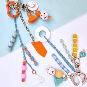 May include: Two decorative keychains featuring gold-tone metal rings and clips. The designs include a variety of colorful beads in orange, pink, yellow, and blue, along with small fabric tassels, white circular charms, and geometric acrylic pendants.