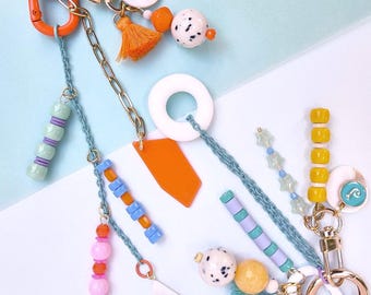 May include: Two decorative keychains featuring gold-tone metal rings and clips. The designs include a variety of colorful beads in orange, pink, yellow, and blue, along with small fabric tassels, white circular charms, and geometric acrylic pendants.