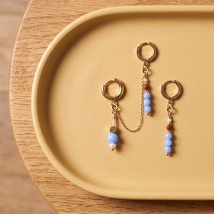 May include: Three gold-tone hoop earrings connected by a delicate gold chain. The earrings feature dangling accents including small light blue spherical beads, tiny white pearls, and small brown textured beads arranged in various vertical patterns.