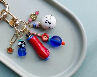 May include: A gold-toned keychain with various colorful charms. Charms include a red and blue cylindrical bead, a white speckled ball, a blue cube, a star, and a flower. The keychain is on a light green tray.