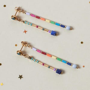 May include: A pair of gold-tone drop earrings featuring two dangling strands each. One strand displays a sequence of multicolored cylindrical beads in lavender, pink, orange, yellow, green, blue, and light blue. The other strand features metallic gold-tone beads, small turquoise accents, and a dark blue square bead at the end.