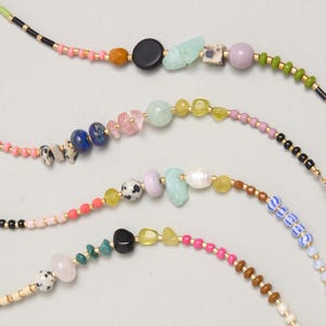 Puede incluir: Cuatro collares de cuentas con una variedad de elementos coloridos, incluyendo pequeñas cuentas de semillas, cuentas redondas lisas, fragmentos de piedra irregulares y cuentas estampadas en tonos rosa, azul, verde, negro, amarillo, marrón y blanco sobre alambre dorado.
