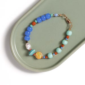 Op de afbeelding: Een kralenarmband met een mix van blauwe, turquoise en oranje kralen, een enkele witte zoetwaterparel en een ronde gele schelpkraal. De armband is afgewerkt met een goudkleurige karabijnsluiting en kleine goudkleurige tussenkralen.