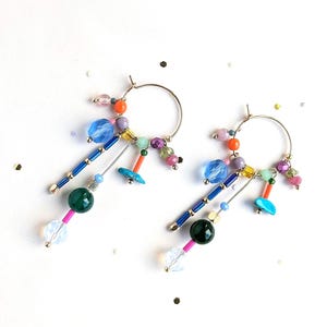 May include: A pair of hoop earrings with colorful beads. The earrings feature gold-tone hoops with dangling strands of blue, green, pink, and clear beads. The earrings have a playful and artistic design.