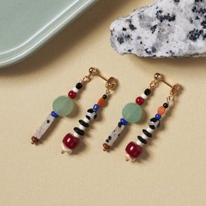 May include: A pair of gold-tone stud earrings featuring dangling multi-colored beads. Each earring has a light green circular bead, a speckled stone bar, and a strand of smaller beads in red, black, white, blue, and orange tones.