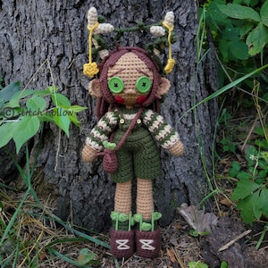 May include: A crocheted doll with brown hair, green eyes, and a green and brown outfit. The doll is wearing a green and brown overalls with a brown satchel and brown shoes. The doll is standing in front of a tree trunk.