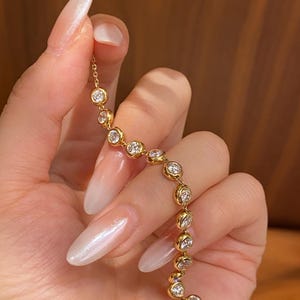 Puede incluir: Una pulsera de cadena dorada que presenta una serie de piedras transparentes redondas engastadas en bisel. Las piedras est&aacute;n unidas en una l&iacute;nea continua, creando un efecto brillante. El metal tiene un acabado de oro amarillo pulido y firme.