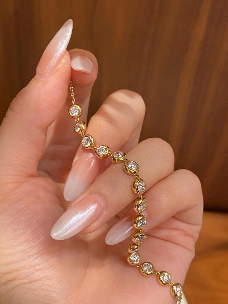 Puede incluir: Una pulsera de cadena dorada que presenta una serie de piedras transparentes redondas engastadas en bisel. Las piedras est&aacute;n unidas en una l&iacute;nea continua, creando un efecto brillante. El metal tiene un acabado de oro amarillo pulido y firme.