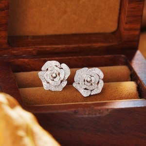 Puede incluir: Un par de pendientes de diamantes en forma de rosa en una caja de madera. Los pendientes están hechos de metal blanco y tienen un diseño delicado.