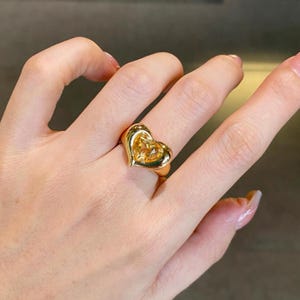 Puede incluir: Un anillo de tono dorado con un engaste prominente en forma de corazón. El centro del corazón contiene una piedra preciosa amarilla translúcida y facetada. La banda y el marco en forma de corazón están hechos de un material metálico pulido.