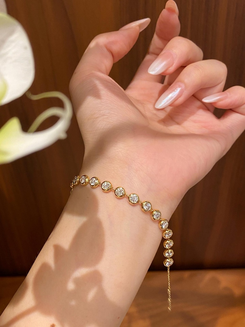 Puede incluir: Una pulsera de tono dorado que presenta una serie de piedras redondas y transparentes engastadas en bisel, unidas en una cadena continua. La pulsera se lleva en la mu&ntilde;eca, mostrando la calidad reflectante de las piedras y los engastes pulidos.