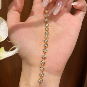 Puede incluir: Una pulsera de cadena dorada con una serie de piedras claras redondas engastadas en bisel dispuestas en un patr&oacute;n lineal. La pulsera consta de catorce engastes circulares individuales, cada uno con una piedra brillante, conectados por una fina cadena dorada.
