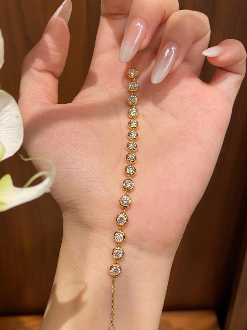 Puede incluir: Una pulsera de cadena dorada con una serie de piedras claras redondas engastadas en bisel dispuestas en un patr&oacute;n lineal. La pulsera consta de catorce engastes circulares individuales, cada uno con una piedra brillante, conectados por una fina cadena dorada.
