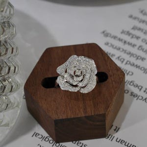 Puede incluir: Un anillo de plata con un diseño de rosa hecho de pequeños diamantes. El anillo se muestra en un soporte de anillo hexagonal de madera.