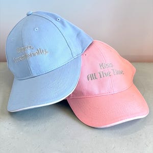 Peut inclure: Deux casquettes de baseball superposées, une bleu clair et une rose. La casquette bleu clair porte la mention brodée "Disco, Occasionally". La casquette rose porte la mention brodée "Kiss All The Time."