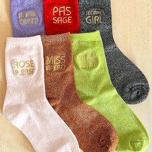 Peut inclure: Six paires de chaussettes colorées avec du texte brodé en or. Les chaussettes sont dans les tons de violet, rouge, noir, rose, marron et vert. Chaque chaussette porte une phrase différente brodée, dont "PAS SAGE" et "MISS APERO".