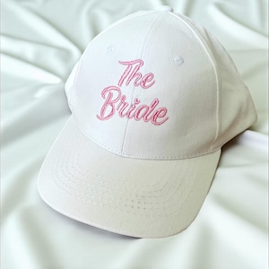 Peut inclure: Casquette de baseball blanche avec une visière incurvée. L'inscription "The Bride" est brodée en rose sur le devant. La casquette est en tissu doux.