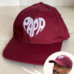 Peut inclure: Une casquette de baseball bordeaux avec une forme de cœur blanche brodée sur le panneau avant. À l'intérieur du cœur, le texte PAPP est cousu dans une police stylisée. La casquette possède une visière incurvée et un bouton sur le dessus.