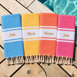 Op de afbeelding: Vier rechthoekige katoenen handdoeken in blauw, geel, oranje en roze, elk voorzien van een brede witte horizontale streep met de namen Julie, Marie, Maman en Julie geborduurd in gouddraad, afgewerkt met decoratieve crèmekleurige franjes.