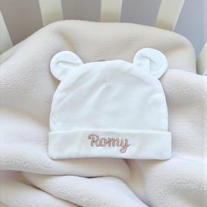 Peut inclure: Un bonnet de bébé blanc avec des oreilles d'ours, brodé du nom "Romy" en fil marron clair. Le chapeau est posé sur une couverture crème, à l'intérieur d'un berceau.