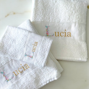 Peut inclure: Un ensemble de trois serviettes en éponge blanche avec le prénom Lucia brodé en fil doré. La lettre L est ornée d'un motif floral coloré. Chaque serviette présente une bordure texturée et une texture en tissu doux et absorbant.