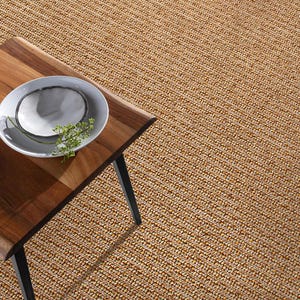 May include: A woven jute rug with a natural, light brown color fills the frame. A wooden table with a live edge and black legs is in the foreground, with a white plate and greenery on top. The rug's texture is visible.