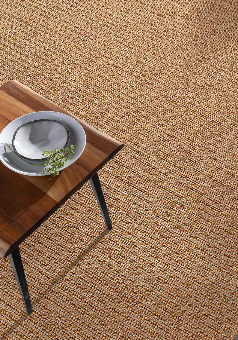 May include: A woven jute rug with a natural, light brown color fills the frame. A wooden table with a live edge and black legs is in the foreground, with a white plate and greenery on top. The rug's texture is visible.