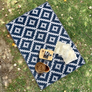 May include: A blue and white geometric patterned picnic blanket with two white pillows, a book, a cup of coffee, and a hat on top of it.