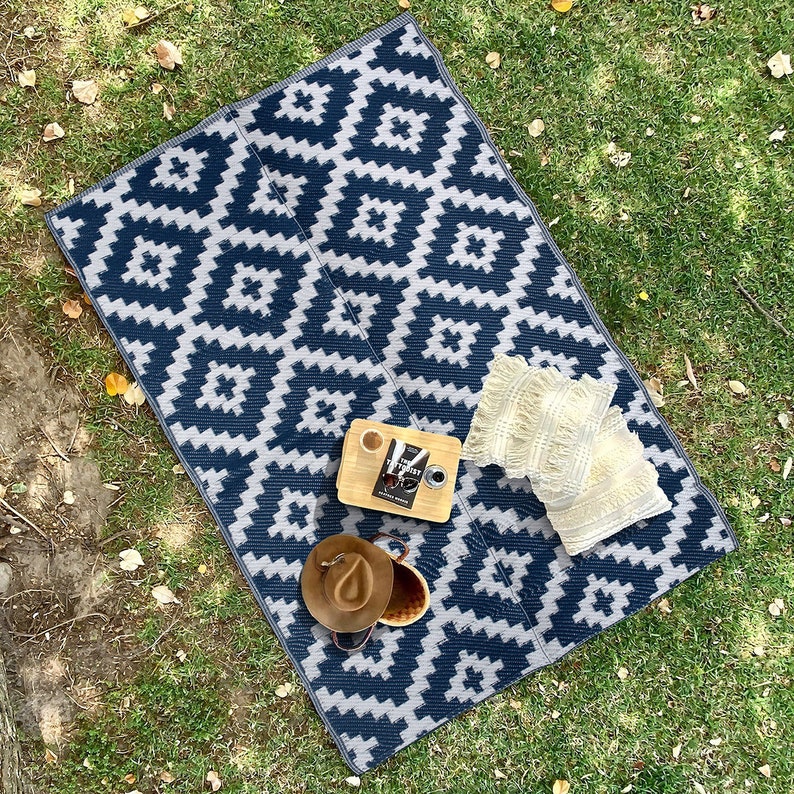 May include: A blue and white geometric patterned picnic blanket with two white pillows, a book, a cup of coffee, and a hat on top of it.