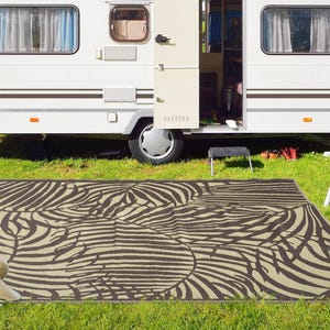 May include: A brown and white patterned outdoor rug with a leaf design. The rug is laid out on green grass in front of a white camper.