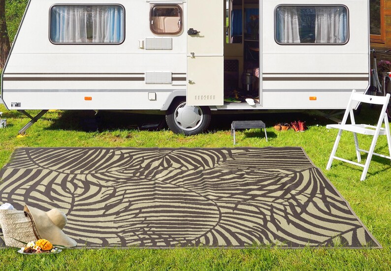 May include: A brown and white patterned outdoor rug with a leaf design. The rug is laid out on green grass in front of a white camper.