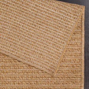 May include: A close-up of a natural fiber rug with a woven texture. The rug is a light tan color with a subtle pattern. The rug is folded over, revealing the underside. The background is a dark wood surface.
