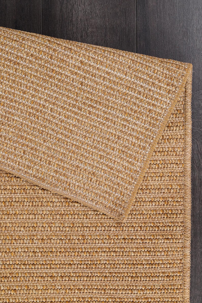 May include: A close-up of a natural fiber rug with a woven texture. The rug is a light tan color with a subtle pattern. The rug is folded over, revealing the underside. The background is a dark wood surface.