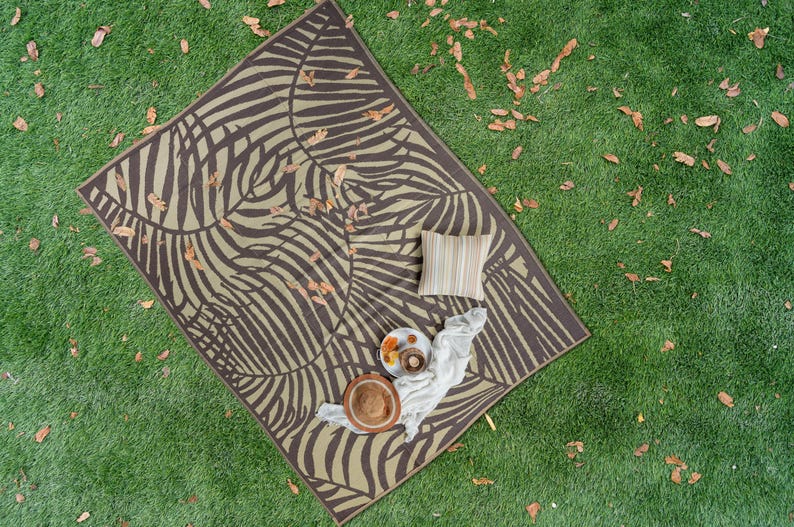 May include: A brown and beige picnic blanket with a zebra stripe pattern. The blanket is laid out on green grass with fallen leaves. There are two plates, a bowl, and a pillow on the blanket.