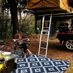 May include: A child sits in a camping chair on a blue and white patterned outdoor rug. A ladder leads up to a pop-up tent on top of a vehicle.