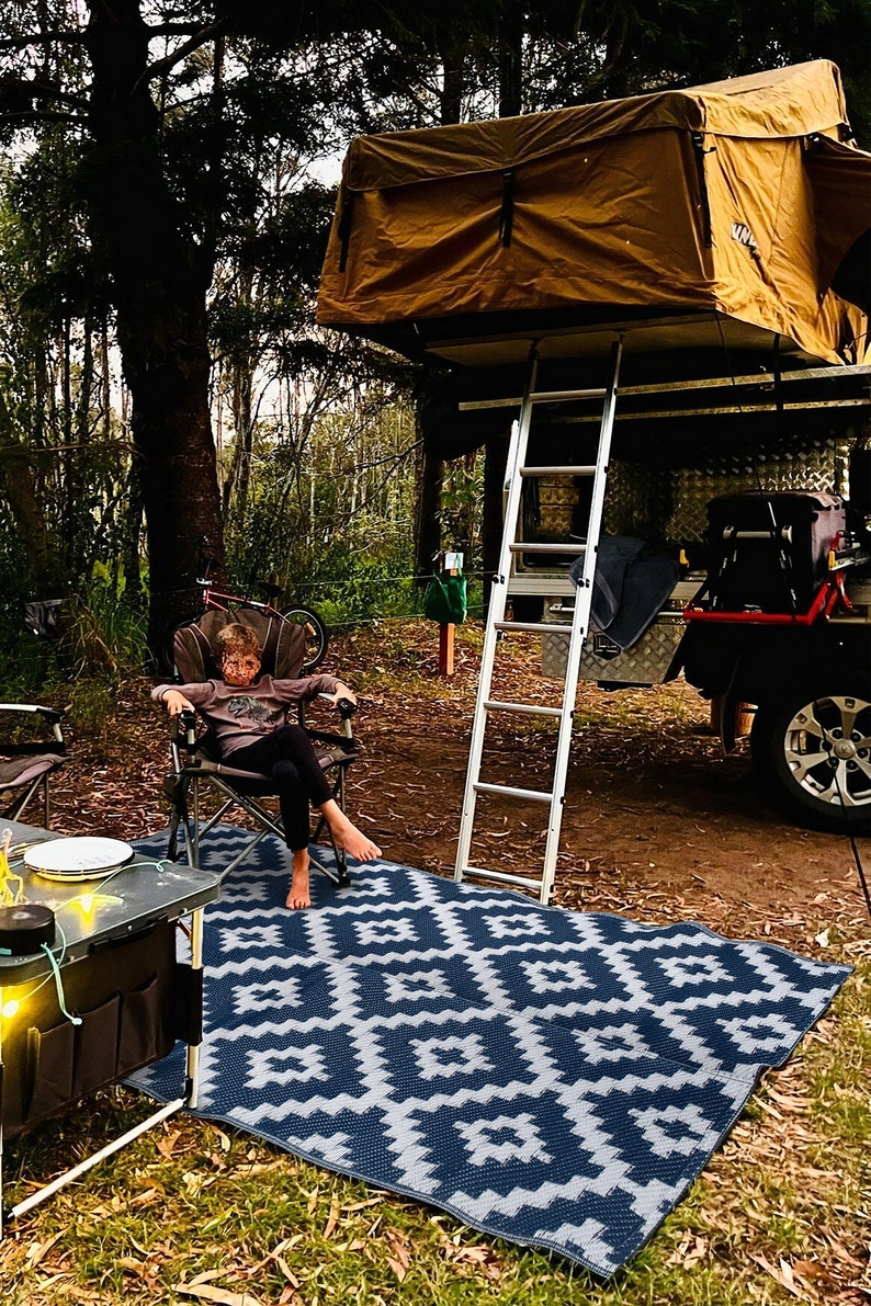May include: A child sits in a camping chair on a blue and white patterned outdoor rug. A ladder leads up to a pop-up tent on top of a vehicle.