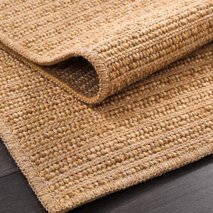 May include: A natural fiber rug with a rolled edge. The rug is made of woven, light brown fibers, with a textured surface and a border. The rug is partially rolled up, revealing the underside and the woven construction.